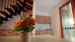 A dining table with a woven basket vase containing flowers, a staircase in the background, and a kitchen area. - Holiday apartment Terraced house with private garden (Rosolina Mare)