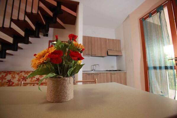 A dining table with a woven basket vase containing flowers, a staircase in the background, and a kitchen area.