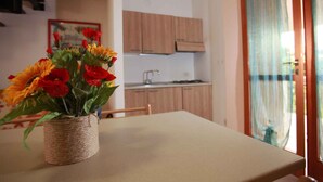 A dining table with a woven basket of flowers, a kitchen with wooden cabinets, and a view of a balcony with a curtain. - Holiday apartment Terraced house with private garden (Rosolina Mare)