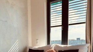 A bedroom with a bed, a window with blinds, and a view of the city.