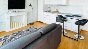 A modern living room with a grey sofa, a flat-screen TV, a fireplace, and a kitchen area with white cabinets and a bar stool.