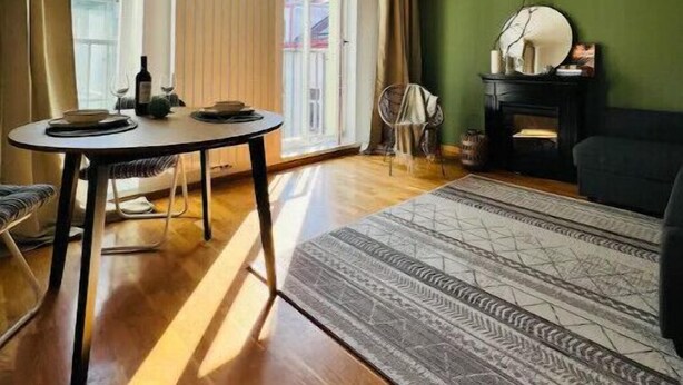 Un salon doté d’un parquet, d’une table à manger ronde entourée de chaises, d’un mur vert, d’une cheminée et d’une grande fenêtre.