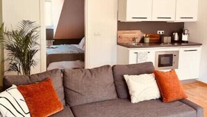 Un salon moderne avec un canapé gris, un petit coin cuisine et une porte-fenêtre donnant sur une chambre.
