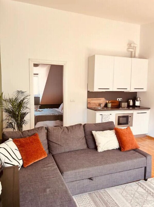 A modern living room with a grey sofa, a small kitchen area, and a doorway leading to a bedroom.