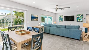 A modern living room with a blue sofa, wooden dining table, and chairs. There is a ceiling fan, a TV mounted on the wall, and artwork on the walls. - Amazing Home on the Intracoastal Waterway! (Indian Rocks Beach)