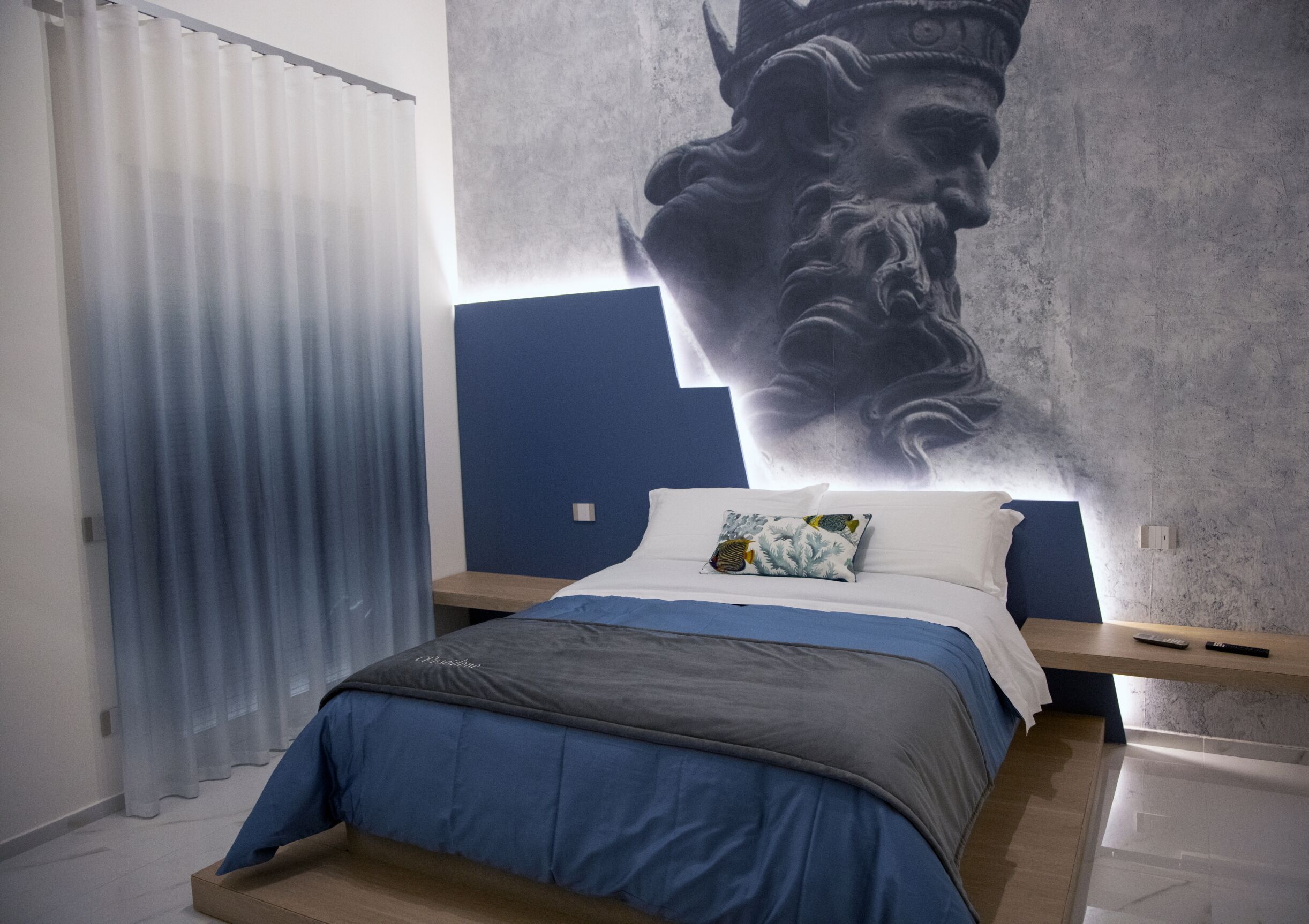 A bedroom with a bed, a nightstand, a large wall mural, and a window with curtains.