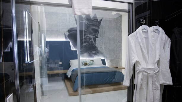 A modern bedroom with a glass partition, a bed with blue bedding, and a white robe hanging on a rack. - Lauria Luxury Room (Gela)