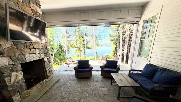 A covered patio with stone flooring, a stone fireplace, wicker furniture, and a view of a lake. - New Maine Private Lake House (Waltham)
