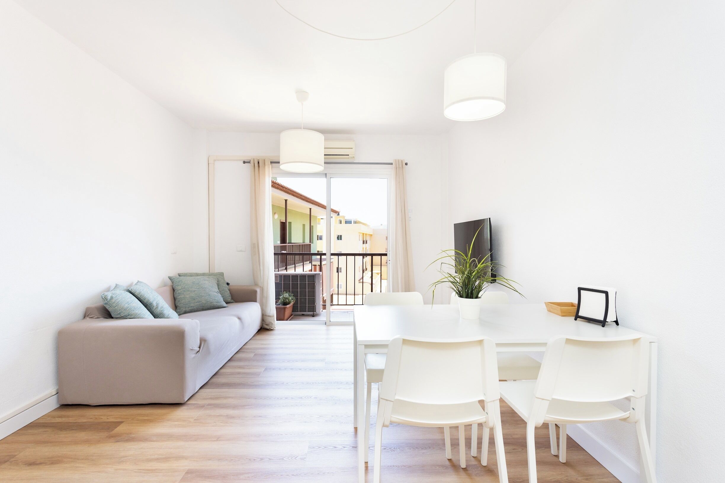 A modern living room with a sofa, dining table, and chairs. There is a balcony visible through the doorway.