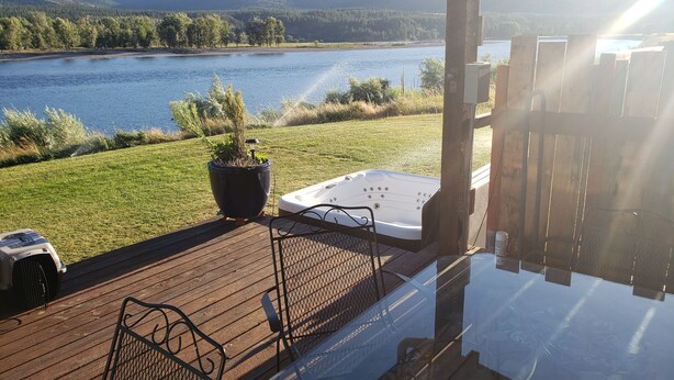 Une terrasse dotée d’un jacuzzi, d’un pot de fleurs et d’un barbecue, offrant une vue sur un lac et des collines boisées.