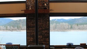 Un salon chaleureux avec une cheminée, des canapés en cuir, une table basse en bois et une grande fenêtre donnant sur un lac et des montagnes.