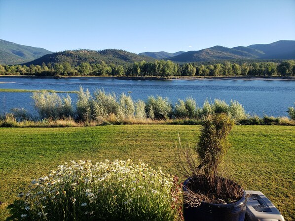 Une vue paisible sur un lac, avec un pré verdoyant au premier plan, un pot de fleurs et une chaîne de montagnes lointaine.