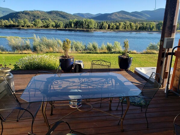 Een terras met een glazen tafel en metalen stoelen met uitzicht op een meer, bomen en heuvels op de achtergrond.
