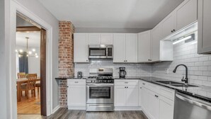 A modern kitchen with white cabinets, a brick accent wall, stainless steel appliances, and a wooden dining area. - Ideal & Exquisite Elmwood Village Victorian (Buffalo)