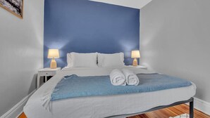 A neatly made bed with a blue blanket and rolled towels, two bedside lamps, and a framed picture on the wall. - Ideal & Exquisite Elmwood Village Victorian (Buffalo)