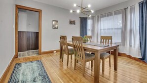 A dining area with a wooden table and chairs, a chandelier, and a view of the outdoors through large windows. - Ideal & Exquisite Elmwood Village Victorian (Buffalo)