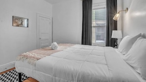 A neatly arranged bedroom with a bed, a bedside lamp, a window with curtains, and a door. - Large Charming Victorian Oasis in Elmwood Village (Buffalo)