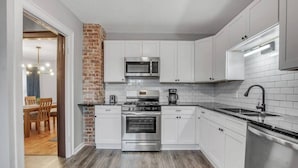 A modern kitchen with white cabinets, a brick accent wall, stainless steel appliances, and a wooden dining area visible through an open door. - Large Charming Victorian Oasis in Elmwood Village (Buffalo)