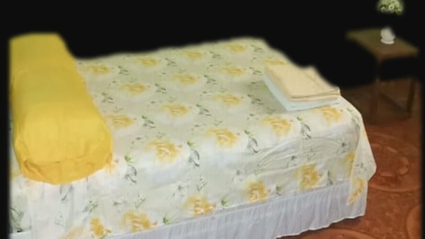 Una cama individual con una colcha estampada con motivos florales, un almohadón amarillo y una mesita de noche con una lámpara.