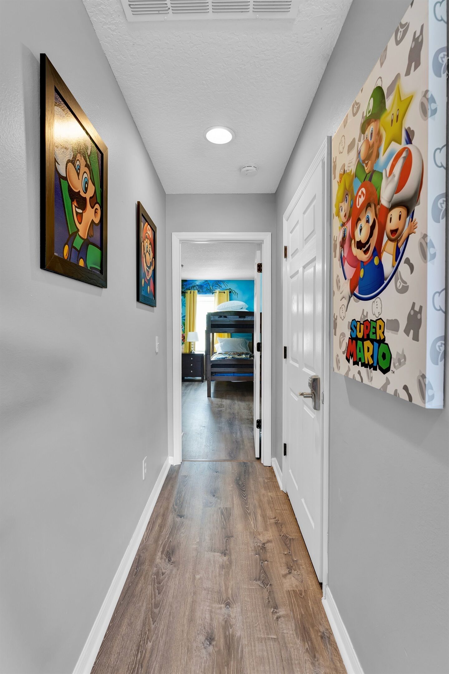 A hallway with framed artwork on the walls, a wooden floor, and a door leading to a bedroom.