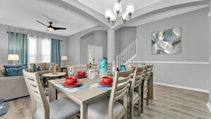 A dining area with a wooden table set for six, surrounded by chairs. A ceiling fan and chandelier are visible above the table.
