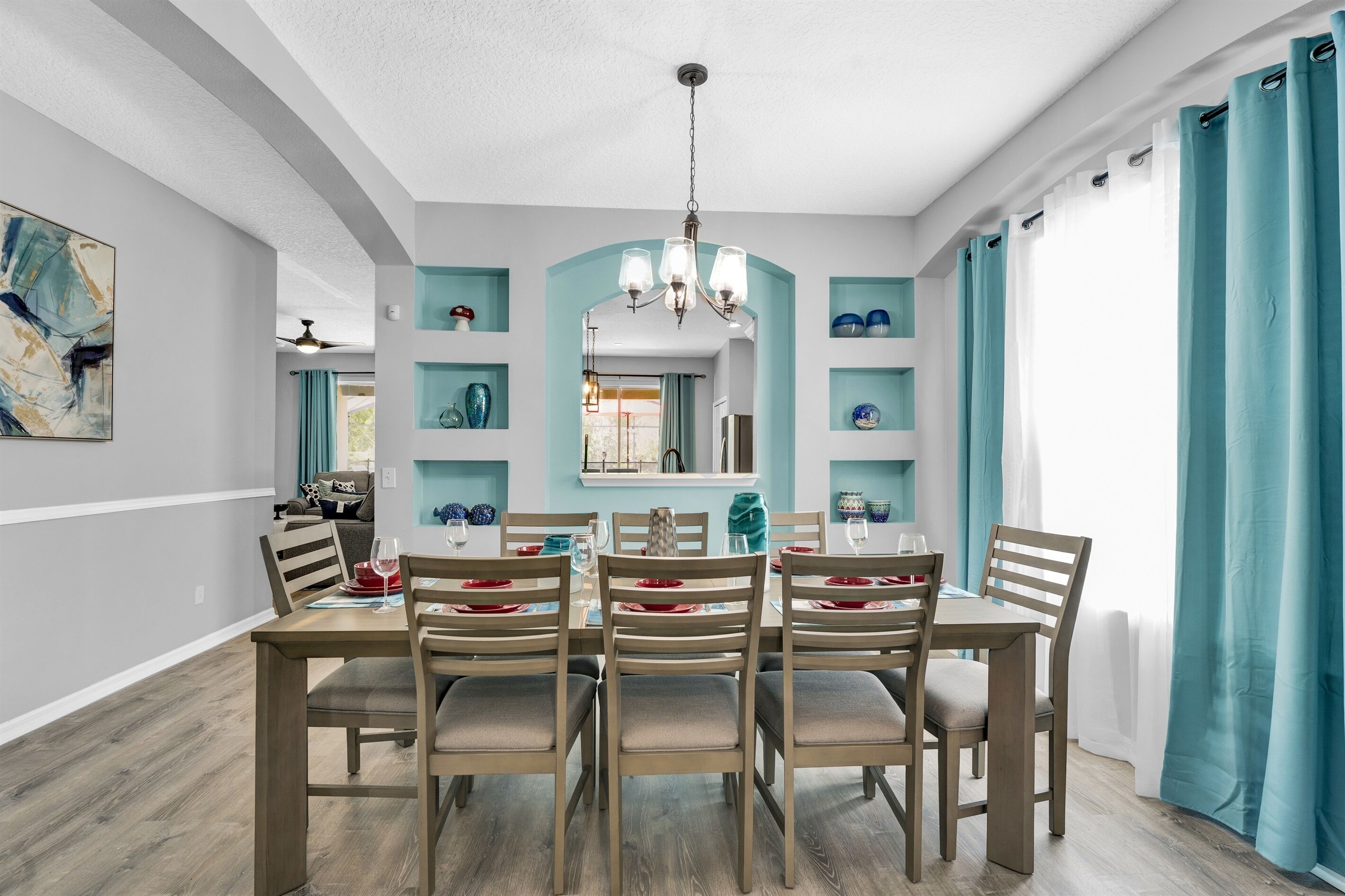 A dining area with a wooden table set for six, light-colored walls, teal shelves, and a chandelier.