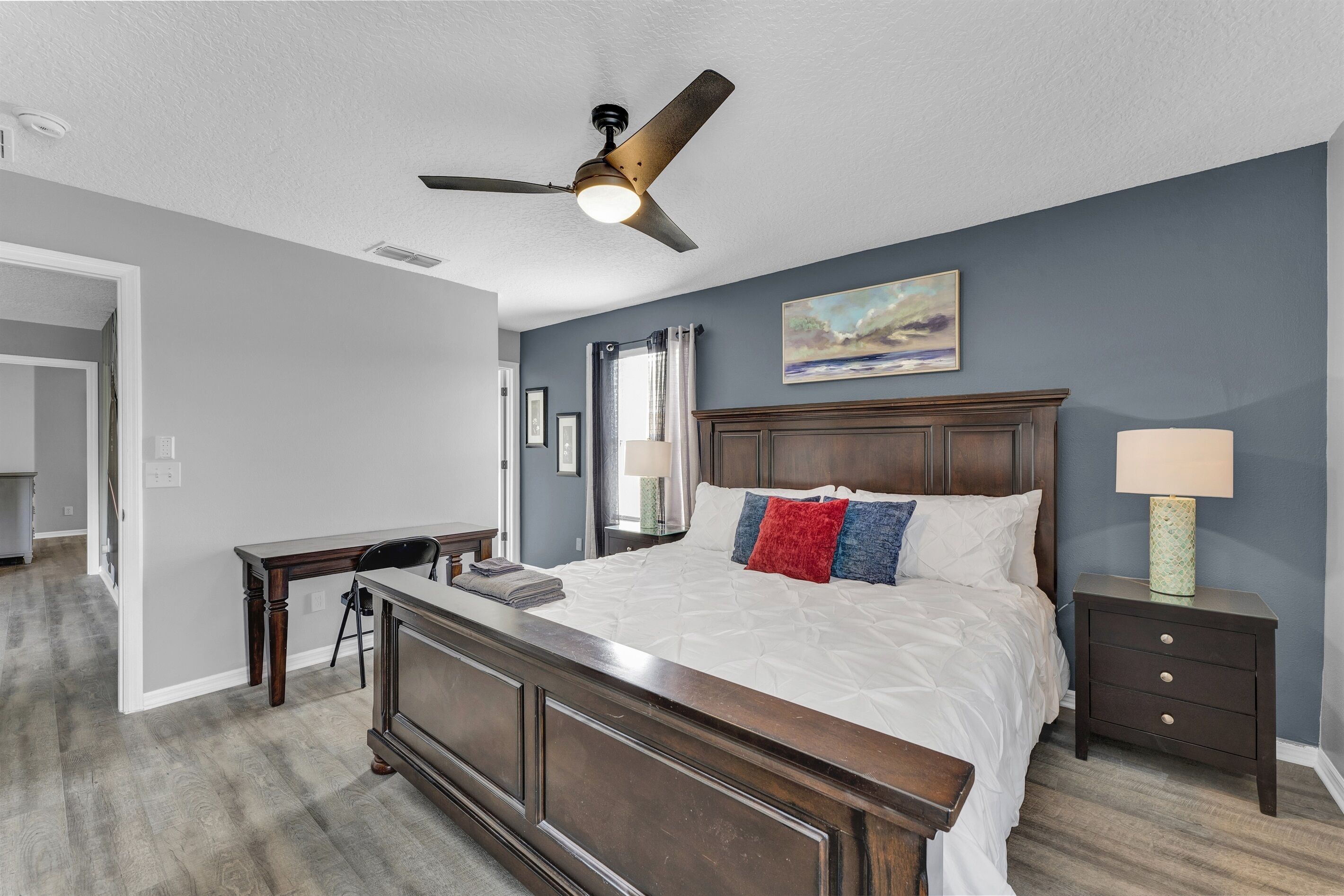 A bedroom with a large bed, a ceiling fan, a nightstand with a lamp, and a painting on the wall.