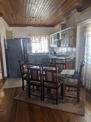 A dining area with a wooden table and chairs, a kitchen in the background, and a ceiling with wooden beams.