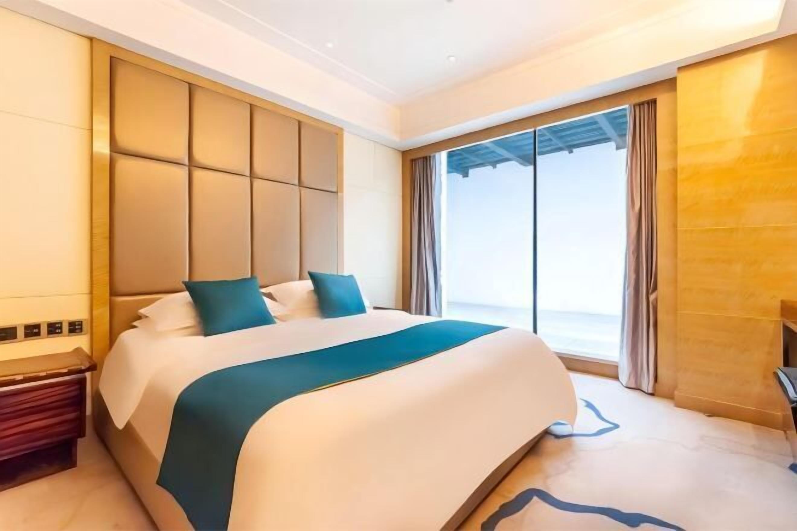 A modern hotel room with a large bed, a built-in headboard, a nightstand, and a large window offering a view.