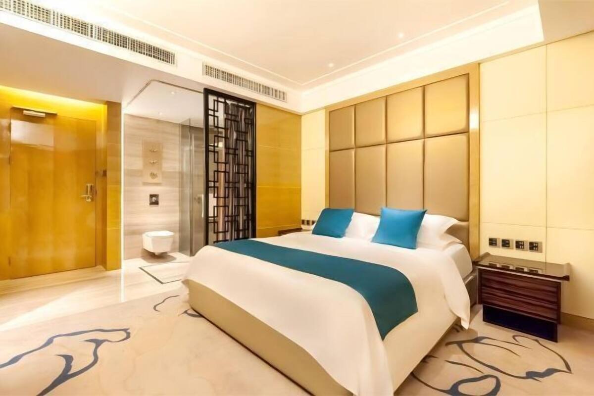 A modern hotel room with a large bed, a wooden wardrobe, a glass partition, and a small bedside table.