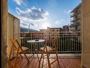 Un balcón con dos sillas de madera y una mesa redonda pequeña, con vista a una zona residencial con edificios y cielo despejado.
