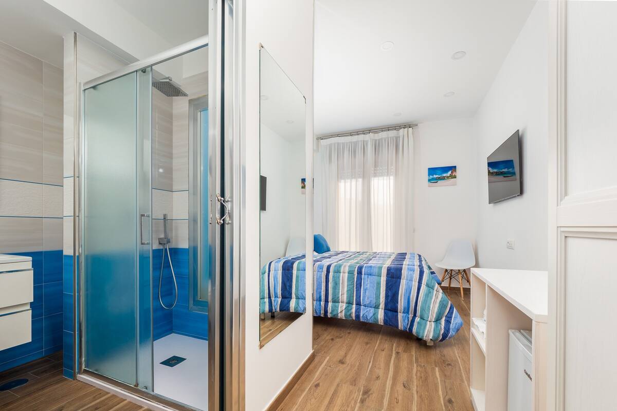 Un hotel moderno con cabina de ducha de vidrio, cama con cubrecama a rayas, escritorio blanco y vista al mar por la ventana.