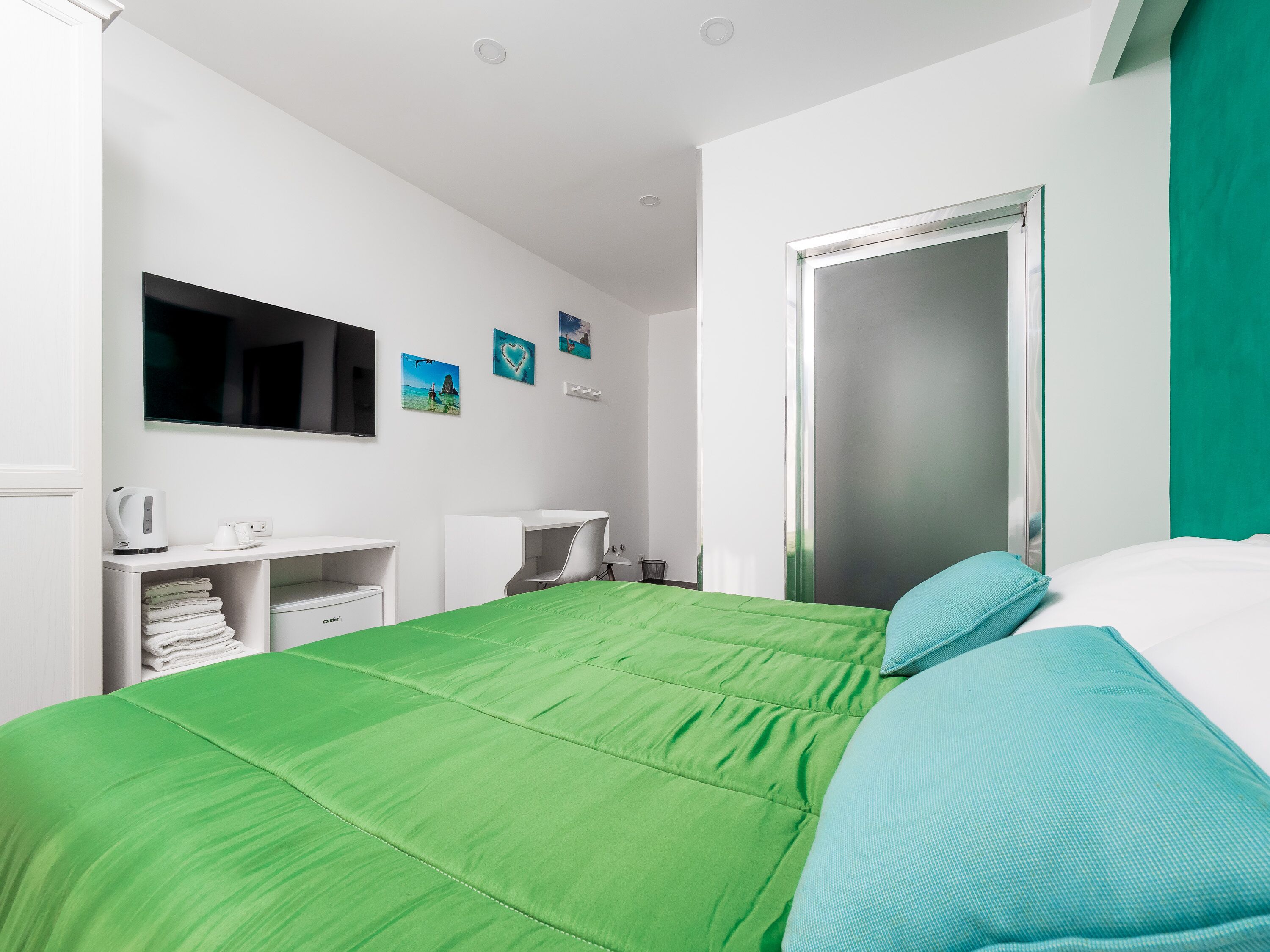 Een moderne hotelkamer met een groen bed, witte muren en een grote spiegel. Er zijn twee nachtkastjes met lampen en een flatscreen televisie bevestigd aan de muur.