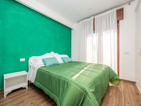 Un dormitorio con pared verde, suelo de madera, cama con ropa de cama verde y mesita de noche blanca.