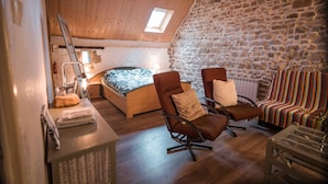 A cozy attic bedroom with a sloped ceiling, a stone wall, a bed with a floral bedspread, two armchairs, and a small table with a lamp.