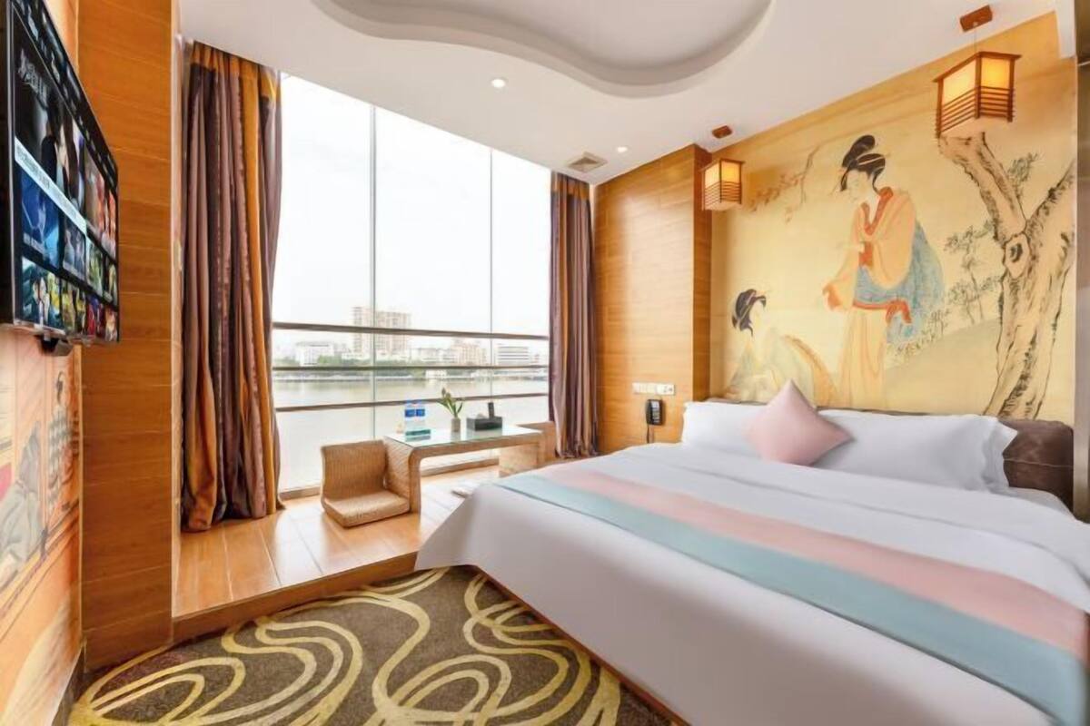 A hotel room with a large bed, a desk, a chair, a view of the city, and traditional artwork on the walls.
