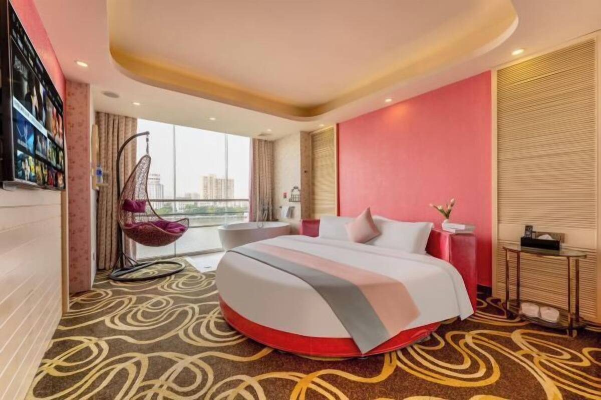 A hotel room with a large bed, a hanging chair, a view of the city, and a red accent wall.