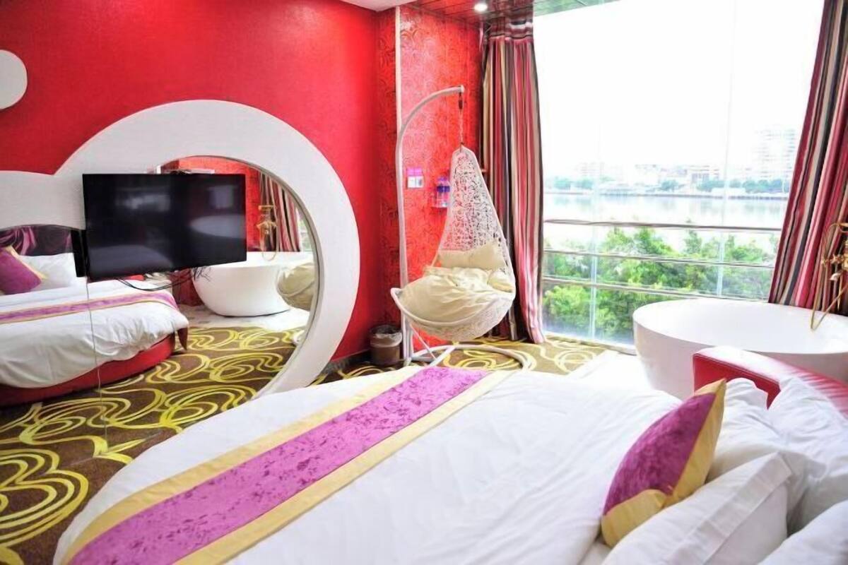 A modern hotel room with a large bed, a TV, a round mirror, a white chair, and a view of the city through large windows.