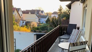 Un balcón con dos sillas blancas y una mesa, con vistas a una zona residencial con casas y árboles.