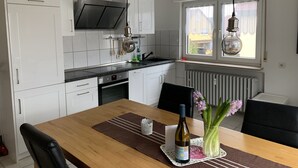 Una cocina moderna con armarios blancos, una encimera negra y una mesa de comedor con sillas. Se ve una ventana con vistas a edificios.