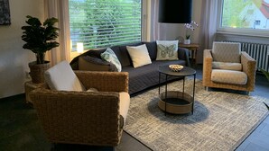 Una sala de estar con un sofá, dos sillones de mimbre, una mesa de centro y un ventanal con persianas.