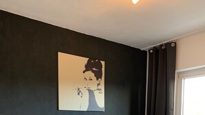 Un dormitorio con cama, mesita de noche, lámpara, ventana con cortinas y un cuadro en la pared.