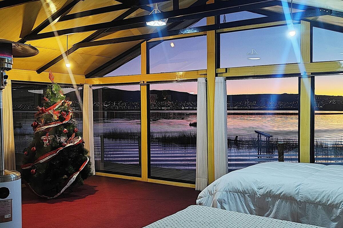 Una habitación con cama, calefacción, un árbol de Navidad y un ventanal con vistas a una masa de agua y al paisaje urbano al anochecer.
