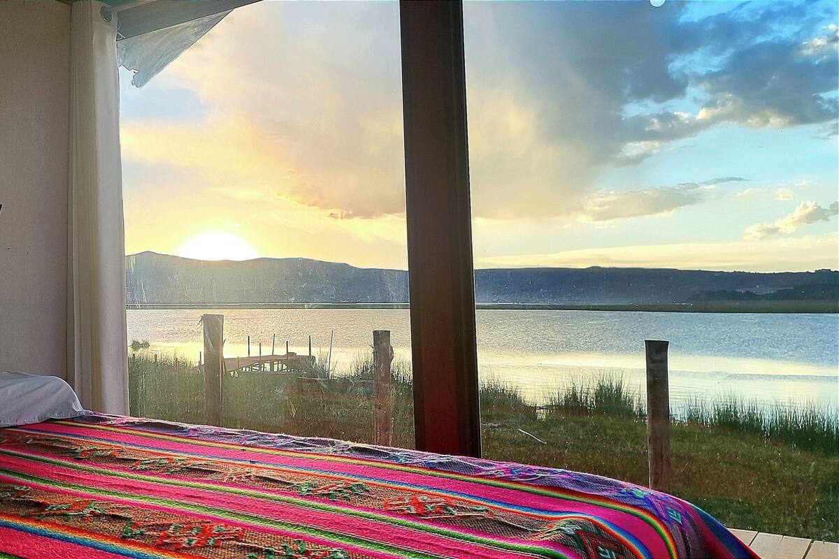 Una habitación con una cama, una manta colorida, una obra de arte de una flor de loto grande y vistas a un atardecer sobre el agua.