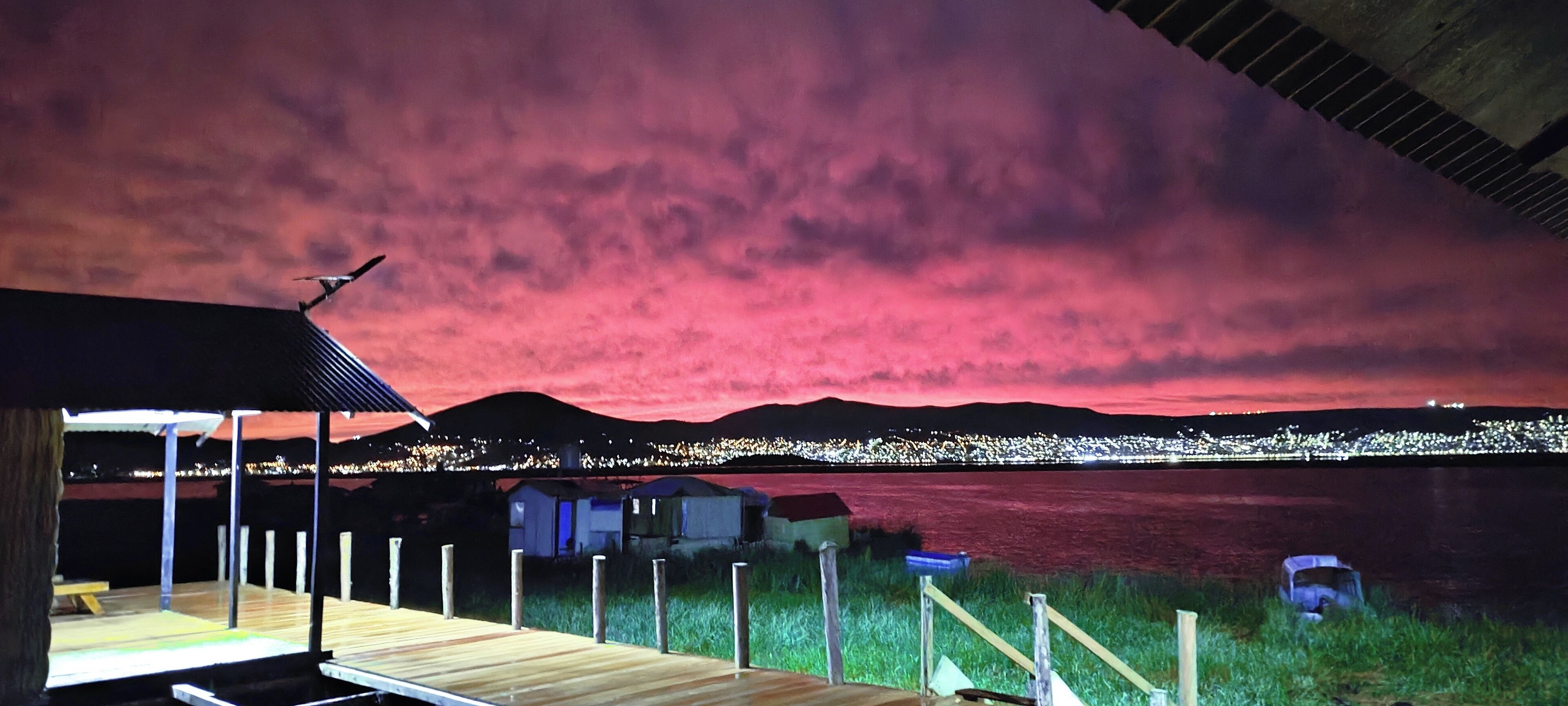 Terraza de madera con vista a un cuerpo de agua, silueta urbana a lo lejos y cielo rosado.