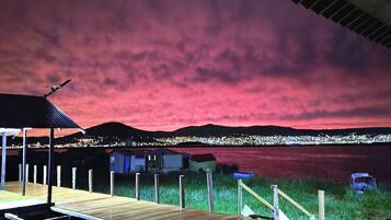 Terraza de madera con vista a un cuerpo de agua, silueta urbana a lo lejos y cielo rosado.