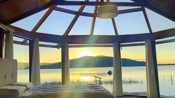 Una stanza con un'ampia finestra a lucernario, un letto e vista su un lago e montagne al tramonto.