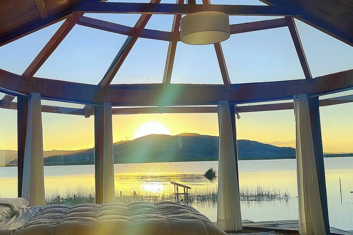 Una habitación con un tragaluz grande, una cama y vistas a un lago y montañas al atardecer.