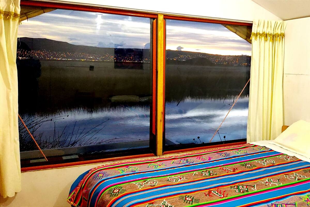 Un dormitorio con un ventanal grande, una cama con manta colorida, y vistas a un lago y luces de la ciudad.