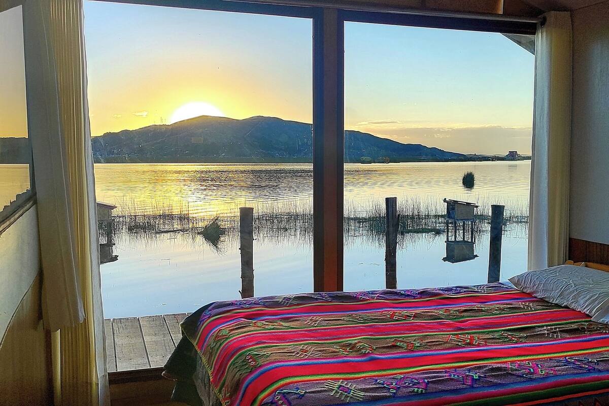 Un dormitorio con un amplio ventanal con vistas a un lago y montañas al atardecer.